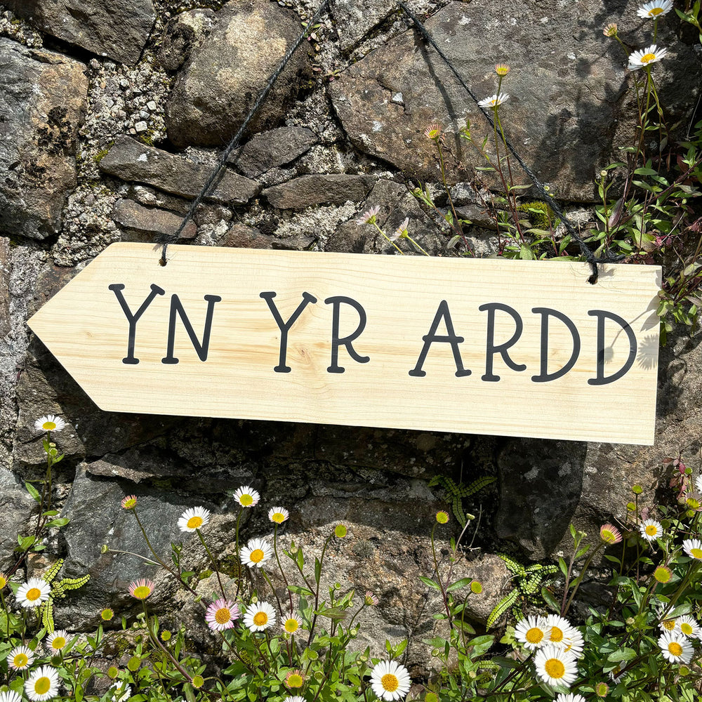 Wooden arrow sign featuring the words 'in the garden' in Welsh - Yn yr ardd and black string for hanging
